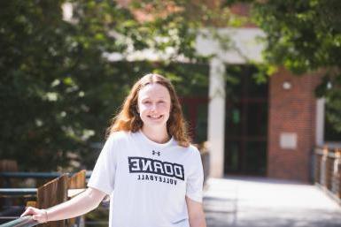 Junior MacKenzie Painter wears a white tshirt that reads "Doane Volleyball." She stands in front of the Lied Science and Mathematics Building.